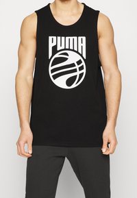 Personne musclée portant un débardeur noir Puma avec un logo de basket-ball blanc, associé à un pantalon noir, debout.