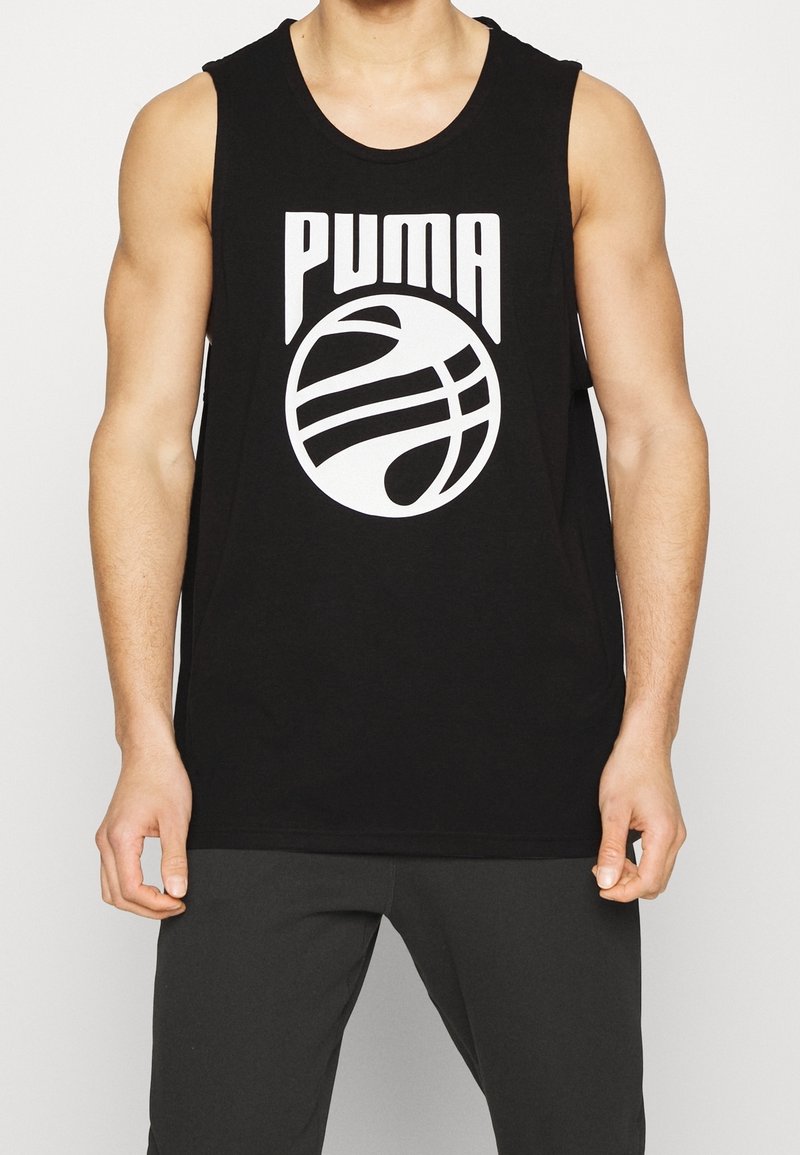 Personne musclée portant un débardeur noir Puma avec un logo de basket-ball blanc, associé à un pantalon noir, debout.