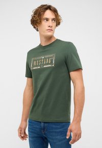 Jeune homme aux cheveux bouclés portant un t-shirt vert avec le logo Mustang et un jean bleu, regardant à sa gauche sur un fond uni.