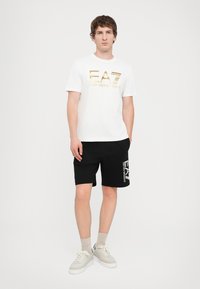 Hvit T-skjorte med gull logo, sorte shorts med hvit logo, lyse sko og lyse sokker. Enkel, uformell atletisk antrekk.