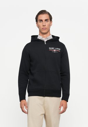 Young man wearing a black zip-up hoodie with "GANT 1949" logo, light striped shirt underneath, and beige pants, standing against a white background.