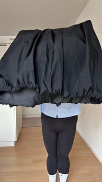 Fekete puffos szoknya, elasztikus derékpánttal ráncolva, sima, fényes textúrával. Fekete leggings fölött viselve.