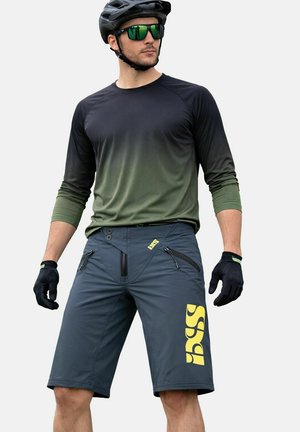 Uomo che indossa una maglia da ciclismo a maniche lunghe nera e verde, pantaloncini grigio scuro con logo giallo, guanti neri, casco e occhiali da sole riflettenti verdi.