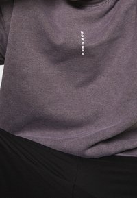 Maglietta grigia in tessuto morbido con scritta verticale bianca "NOWHERE" sul petto. La maglietta ha una vestibilità comoda e scollo arrotondato.