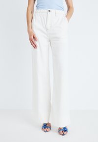 Pantalons large blancs avec taille-haute, fabriqués en tissu lisse. Caractéristiques des poches latérales et une fermeture à bouton. Associés à des sandales bleues.