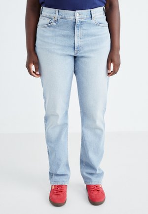 Persoon die lichtblauwe straight-fit jeans en rode sneakers draagt, staand tegen een effen witte achtergrond.