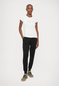 White fitted t-shirt with "ARMANI EXCHANGE" logo in metallic rose gold, paired with black tapered joggers and olive green sneakers.