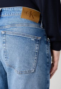 Ljusblå denimjeans med hög midja, som har en brun läderpatch med Calvin Klein Jeans-logotyp och en standarddesign för bakfickor.