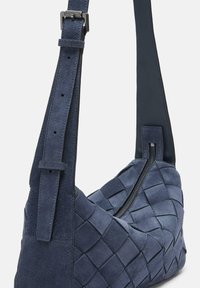 La borsa a tracolla blu in suede presenta una texture intrecciata, una tracolla regolabile e una chiusura con zip, evidenziando un design moderno e minimale.