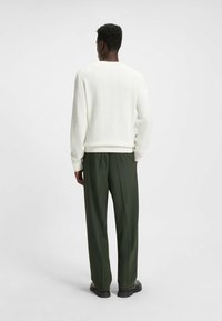 Karamellfarbener Strickpullover mit strukturiertem Muster, kombiniert mit dunkelgrünen, plissierten Hosen. Schwarze Schuhe runden das Outfit ab.