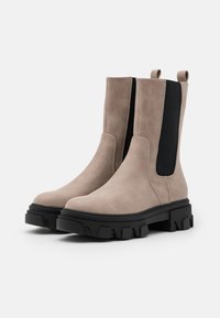 Ett par beige mockaläder Chelsea-boots med svarta elastiska sidopaneler och kraftiga svarta gummisulor, visade mot en vit bakgrund.