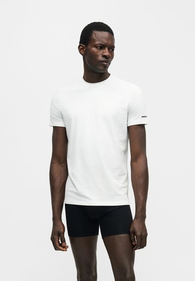 Άνδρας που φοράει απλό λευκό κοντομάνικο μπλουζάκι και μαύρα εφαρμοστά σορτς, στέκεται μπροστά σε λευκό φόντο, κοιτάζοντας αριστερά του.