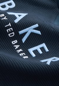 Marineblåt tekstureret stof med store broderede bogstaver i lyseblå. Bogstaverne staver "BAKER" og inkluderer "by TED" i mindre hvid tekst.