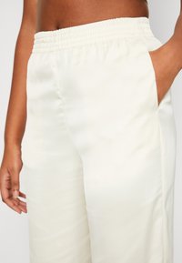 Close-up van een persoon die witte elastische broek met tailleband draagt, met één hand in de zak en de andere hand ontspannen langs het lichaam.