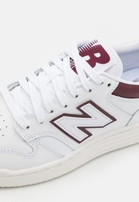 Zapatilla de cuero blanca con acentos en burdeos, presenta cordones blancos, área de los dedos perforada y un prominente logotipo "N" en el costado. Suela texturizada.