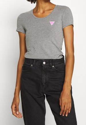 Kvinde iført en tætsiddende grå t-shirt med korte ærmer, med en lille lyserød Guess-logo og højtaljede sorte jeans mod en ensfarvet baggrund.