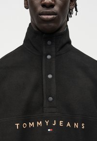 Κοντινό πλάνο ενός ατόμου που φοράει μαύρο φλις πουλόβερ με ψηλό γιακά και τέσσερα κουμπιά, με το κέντημα "TOMMY JEANS" σε μπεζ κάτω από αυτά.