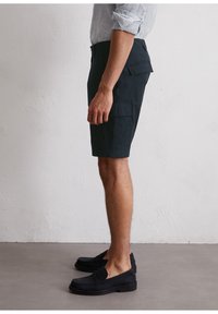 Pantalones cortos de carga navy hechos de una mezcla de algodón, con bolsillos laterales, frente plano y un ajuste relajado, combinados con zapatos negros tipo slip-on.