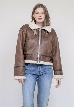 Femme portant une veste aviateur en peau retournée marron avec une fermeture éclair frontale et un jean bleu, debout devant un fond clair uni.