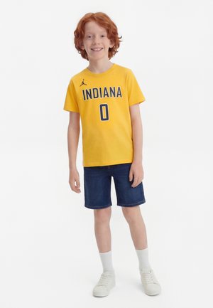 Garçon aux cheveux roux souriant, portant un maillot jaune de l'Indiana avec le numéro 0, un short bleu foncé, des chaussettes blanches et des baskets blanches, debout sur un fond blanc.