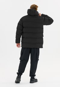 Musta puhvitakkia, jossa on huppu, ominaisuutena tikattu muotoilu ja joustavat hihansuut. Yhdistetty mustiin cargo-housuihin ja urheilukenkiin.