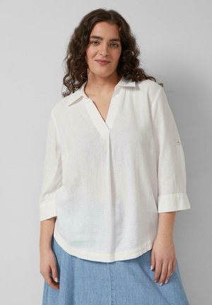 Kobieta o kręconych ciemnych włosach, ubrana w luźną białą bluzkę bez guzików z rękawami do połowy łokcia oraz jasnoniebieską spódnicę jeansową, stoi na tle jednolitego tła.