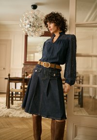 Blouse bleu marine avec manches en dentelle associée à une jupe en jean à poches avant, accentuée par une ceinture marron à boucle circulaire et des bottes hauteur genou.