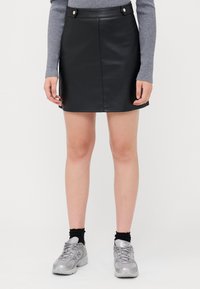 Mini-jupe en simili cuir noir avec une forme droite, dotée de deux boutons argentés à la taille, associée à un haut gris à manches longues et des baskets.