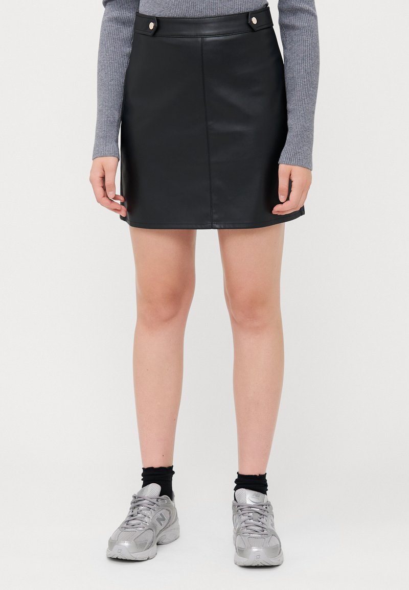 Mini-jupe en simili cuir noir avec une forme droite, dotée de deux boutons argentés à la taille, associée à un haut gris à manches longues et des baskets.