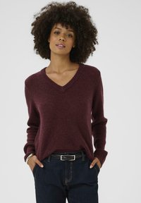 Bordo pulover z V-izrezom iz mehke pletenine, z dolgimi rokavi in sproščenim krojem, v kombinaciji s temnimi jeans hlačami in črnim pasom.
