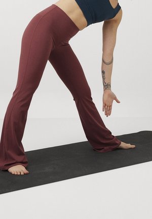 Osoba ubrana w bordowe spodnie do jogi i czarny top rozciąga się na czarnej macie, dotykając palców u stóp lewą, tatuażowaną ręką wyciągniętą w dół.