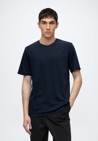 Young man with short dark hair wearing a plain dark blue t-shirt and black pants, standing with one hand in pocket against a white background.