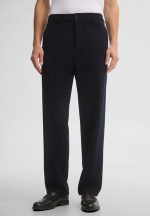 Uomo che indossa pantaloni formali neri, scarpe eleganti nere e una camicia bianca su uno sfondo chiaro e semplice.