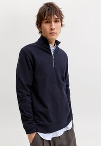 Σκούρο μπλε πουλόβερ με υψηλό γιακά και φερμουάρ μέχρι το μισό μήκος. Διαθέτει ριμπ μανσέτες και τελείωμα, φορεμένο πάνω από μια ελαφρώς γαλάζια πουκάμισο με γιακά.