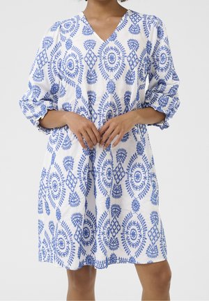 Kvinde iført en hvid knælængde kjole med blå indviklede blomster- og geometriske mønstre, med V-hals og 3/4 puffede ærmer.