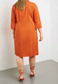 Vestito arancione alla lunghezza del ginocchio con maniche a tre quarti, texture morbida, cuciture laterali e un taglio semplice. Indossato con sandali arancioni con cinturini.