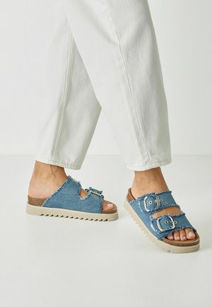 Sandały na płaskiej podeszwie z denimu z dwoma metalowymi klamrami, postrzępionymi krawędziami i beżową gumową podeszwą zygzakowatym wzorem. Otwarty przód.