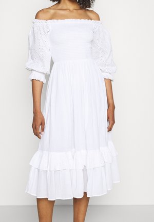 Vrouw draagt een witte off-shoulder midi-jurk met opgezette oogje-mouwen en een geplooide zoom, staand voor een effen achtergrond.