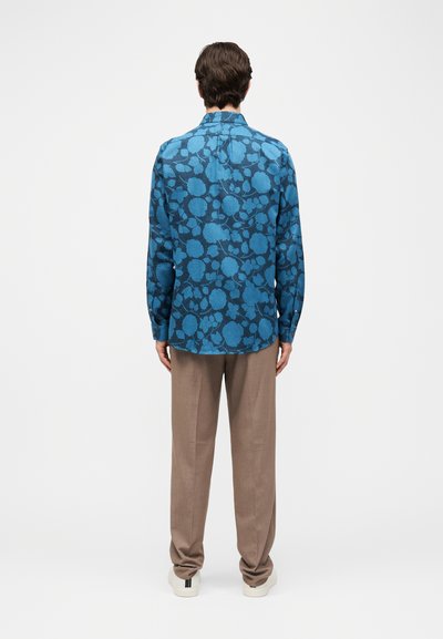 Άντρας που φοράει μπλε πουκάμισο με λουλουδένιο σχέδιο μακριάς μανίκας, μπεζ παντελόνι και λευκά αθλητικά παπούτσια, στέκεται με την πλάτη γυρισμένη σε απλό φόντο.