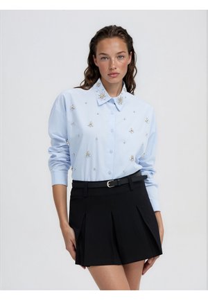Donna che indossa una camicia azzurro chiaro con bottoni e decorazioni in perle e gemme, infilata in una minigonna nera plissettata con cintura.