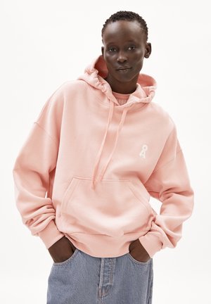 Hoodie rosa con tasca a marsupio, cappuccio con cordino e dettagli del logo in nastro bianco. Indossato con jeans in denim blu, caratterizzati da una vestibilità rilassata.