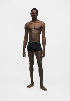 Αθλητικός άνδρας με σκουρόχρωμο δέρμα, όρθιος ξυπόλητος, φορώντας μαύρα εσώρουχα Calvin Klein σε απλό λευκό φόντο.