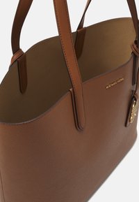 Hnědá kožená taška přes rameno s texturovaným povrchem, hladkými uchy a zlatým logem. Design s otevřeným vrchem a prostorným interiérem.