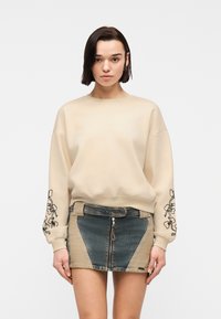 Γυναίκα που φοράει μπεζ φούτερ με μαύρη κεντημένη λουλουδένια μανσέτα και δίχρωμη μίνι φούστα τζιν με φερμουάρ μπροστά, με λευκό φόντο.