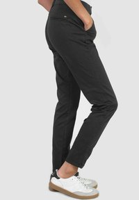 Zwarte slim-fit broek met een gladde textuur, voorzien van zijzakken en een achterzak met rits, gecombineerd met witte sneakers en een grijze achtergrond.