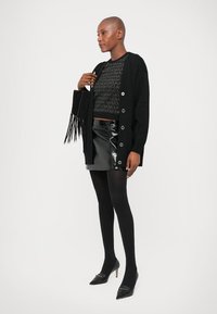 Kvinne i svart mønstrete genser, lang svart cardigan, glitrende svart miniskjørt, svarte strømpebukser, svarte hæler og holder en svart frynseveske.