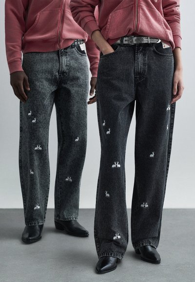 Zwart-grijze spijkerbroek met witte geborduurde konijn ontwerpen. Losse pasvorm, rechte pijpen, gedragen met zwarte puntige laarzen en een roze hoodie.