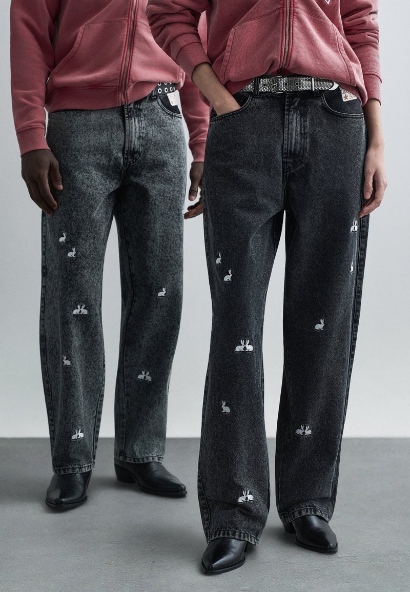 Svart- och gråa jeans i denim med vita broderade kanindesigner. Lös passform, raka ben, kombinerade med svarta spetsiga stövlar och en rosa hoodie.