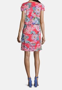 Šatové šaty s květinovým potiskem a vrstevnatým designem v odstínech růžové, doplněné modrými sandály na vysokém podpatku. Průhledná látka má živé květiny.