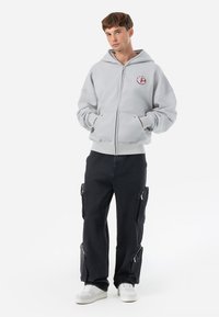 Light gray zip-up hoodie with front pockets and a small logo on the left chest, paired with black cargo pants featuring zippered pockets.